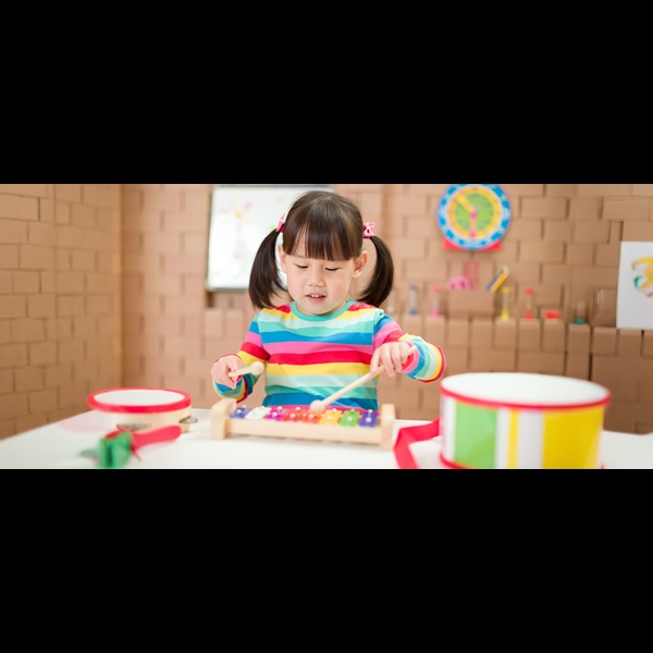 Young girl in a bright striped jumper playing a coloured xylophone, surrounded by musical instruments.