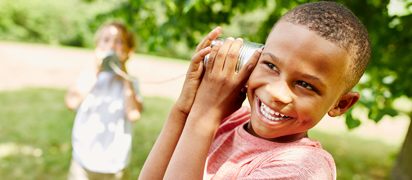 Two children playing with a tin-can telephone outside in a park. 