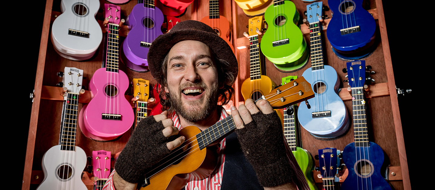Man in fingerless gloves and hat, smiling and playing the ukulele, in front of brightly coloured ukuleles. 