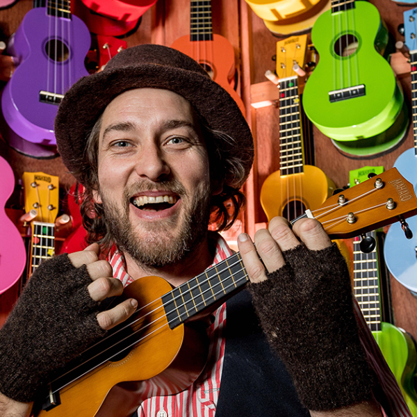 Man in fingerless gloves and hat, smiling and playing the ukulele, in front of brightly coloured ukuleles.