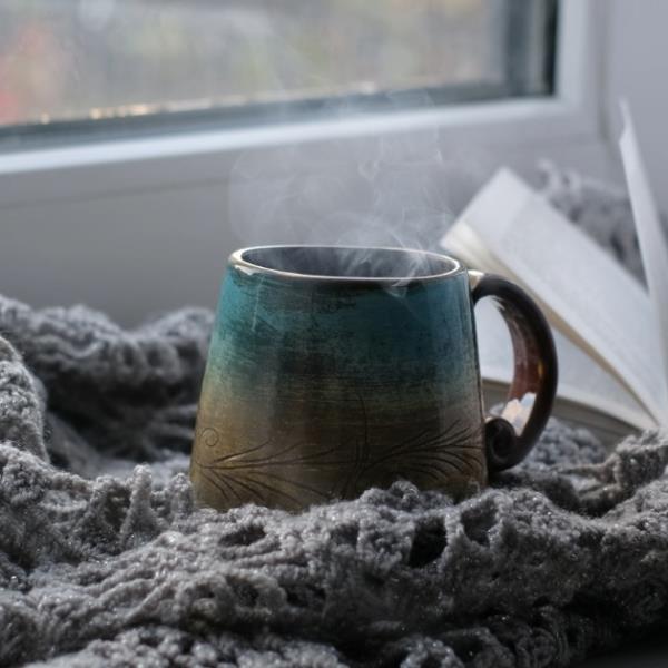 Steaming hot drink and book, resting on a grey blanket by a window with winter weather outside.