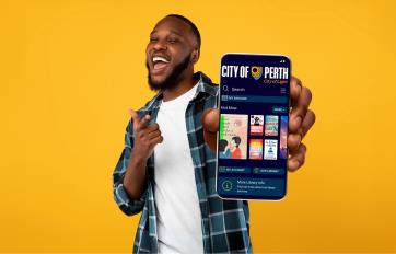 Handsome, cheerful man smiling into the camera and holding out his phone showing the screen with the City of Perth Library app on it.